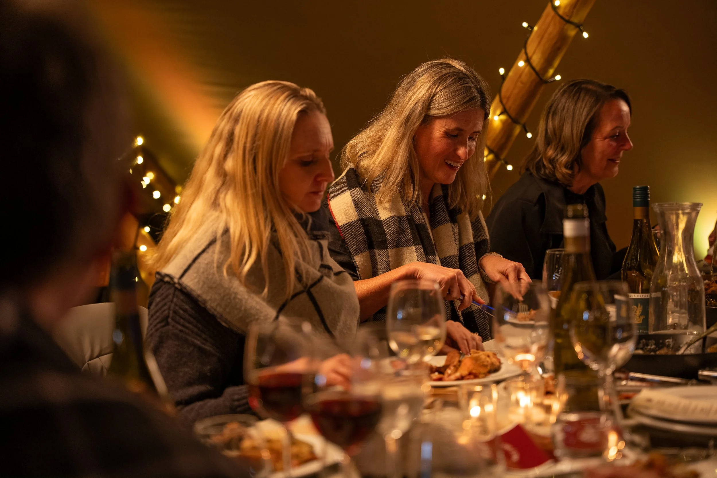 Women dining at a festive gathering with candles, wine, and string lights.