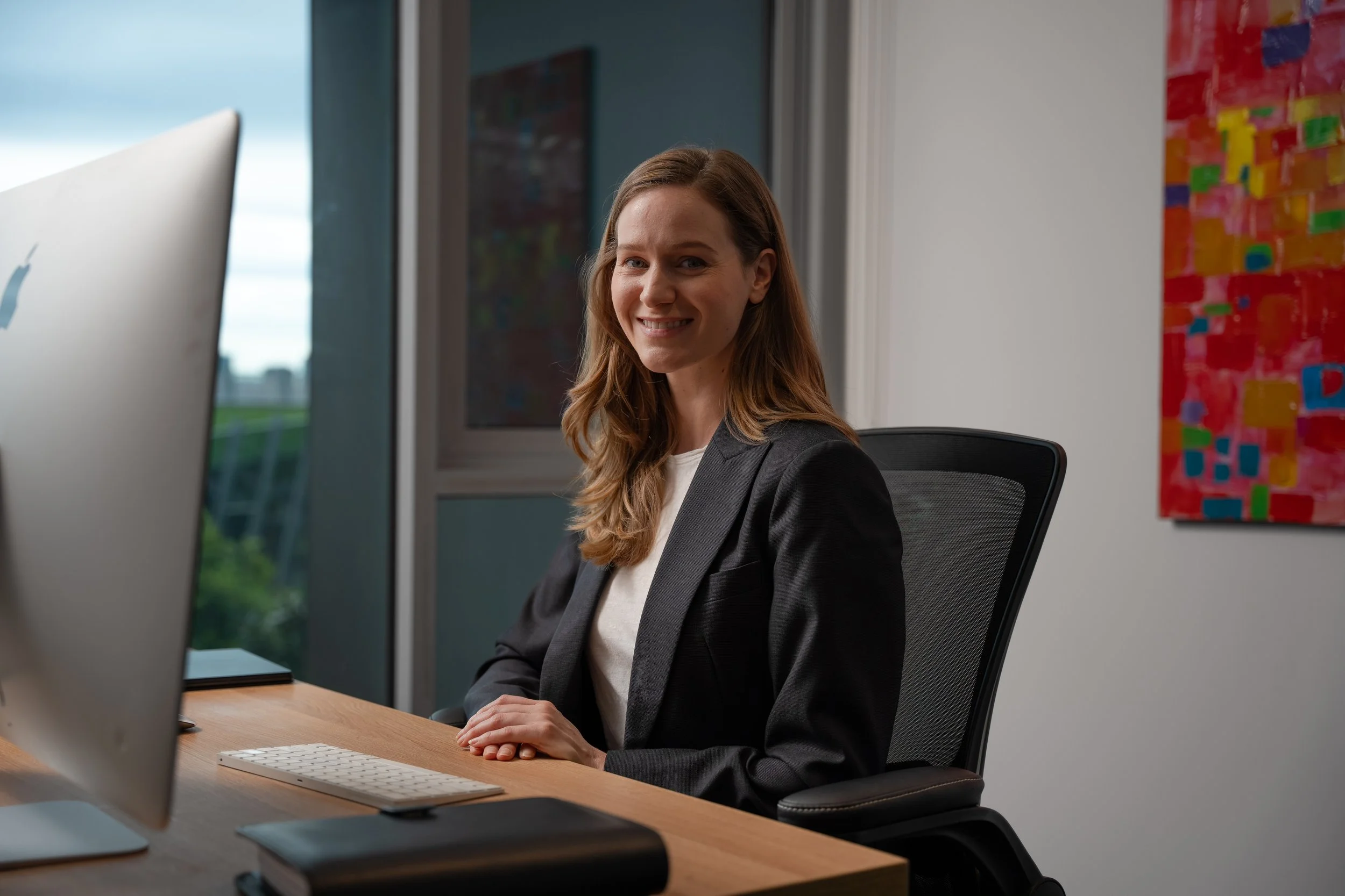 A woman with shoulder-length red hair wearing a black blazer sitting at a desk in front of a computer in an office.