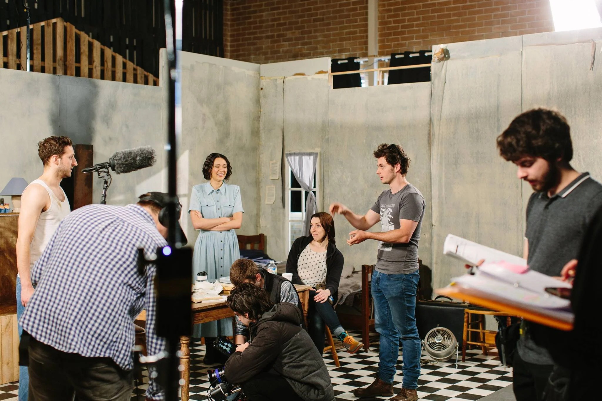Scene from a film or TV show set, featuring multiple crew members and actors in a room with unfinished walls, a checkered floor, and filming equipment.