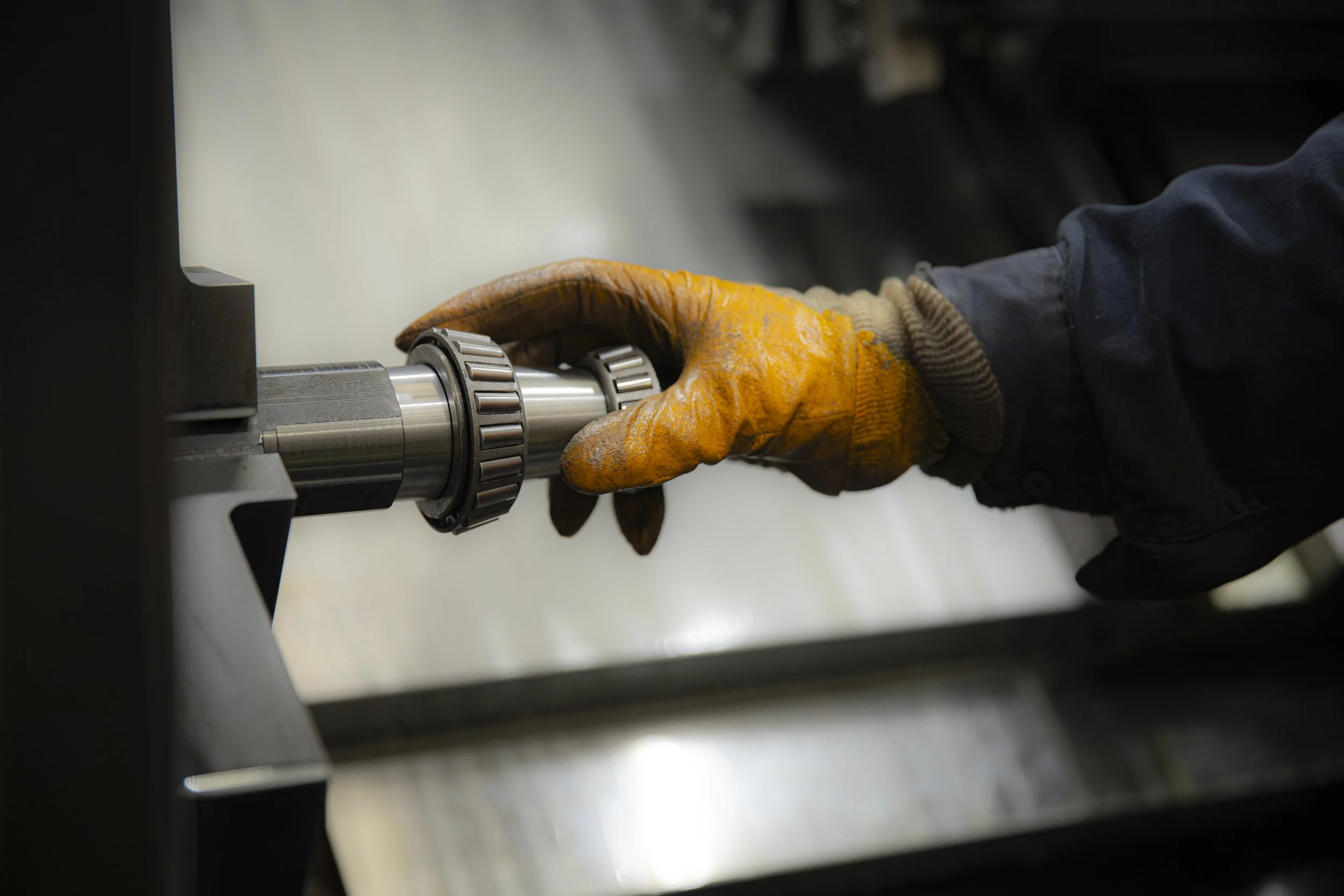 A worker's hand wearing a yellow glove is holding and inspecting a shiny cylindrical metal shaft with a textured ring around it, positioned horizontally on a metallic surface.