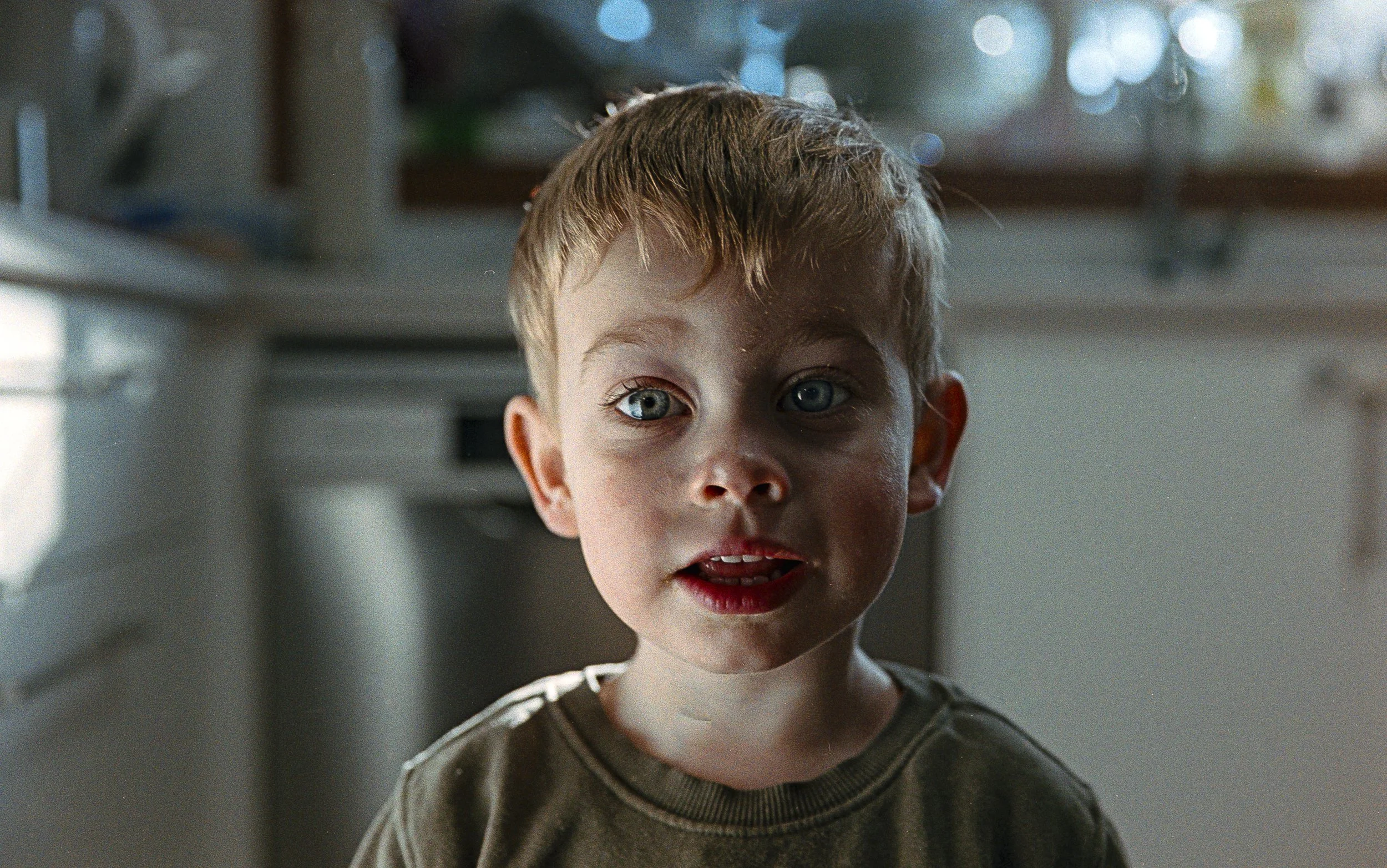 A young boy with blond hair and blue eyes looking directly at the camera, standing in a kitchen with sunlight coming through the window in the background.