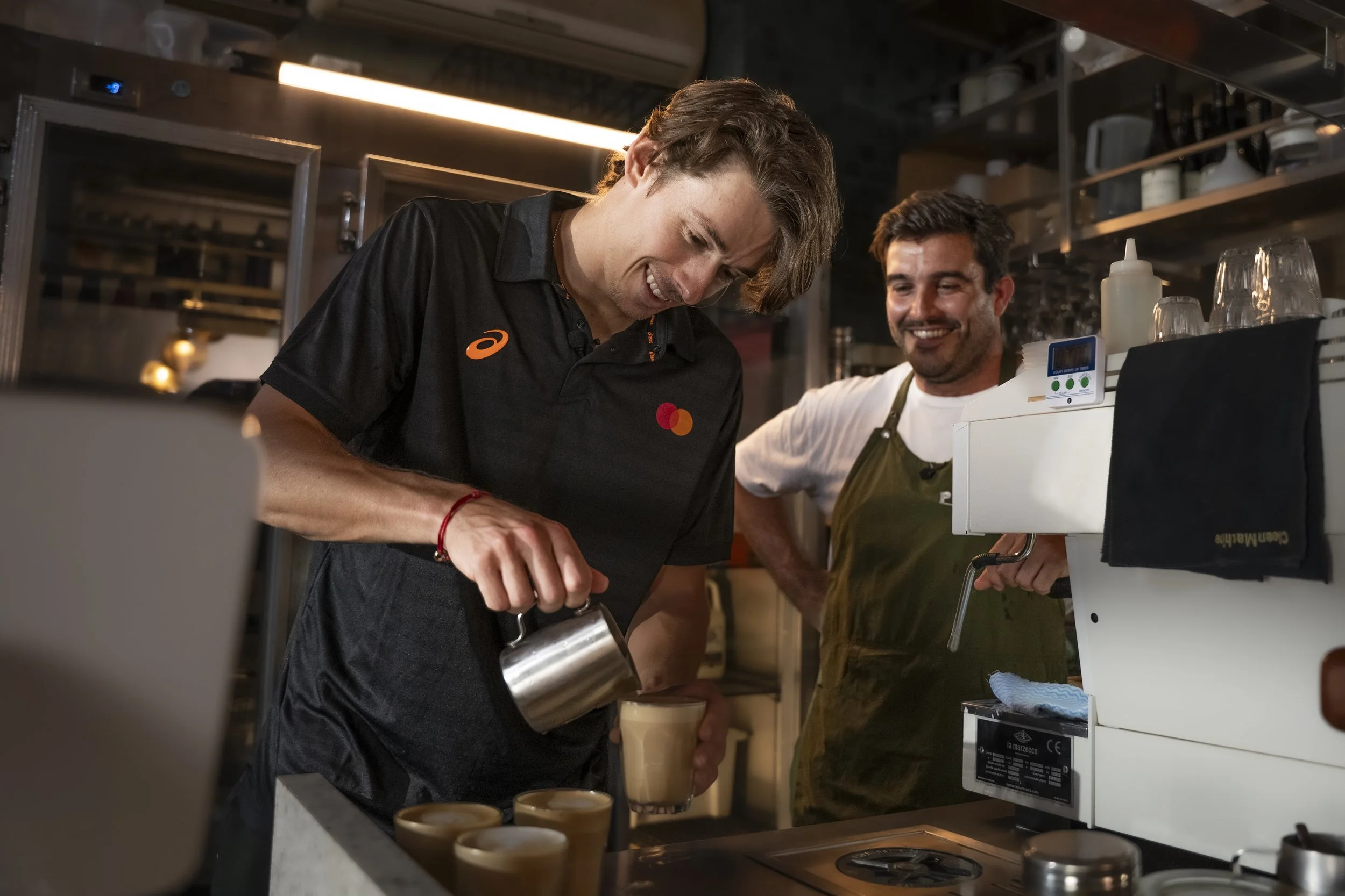 Two men working in a coffee shop, one pouring a drink and the other smiling, surrounded by coffee-making equipment and glasses.
