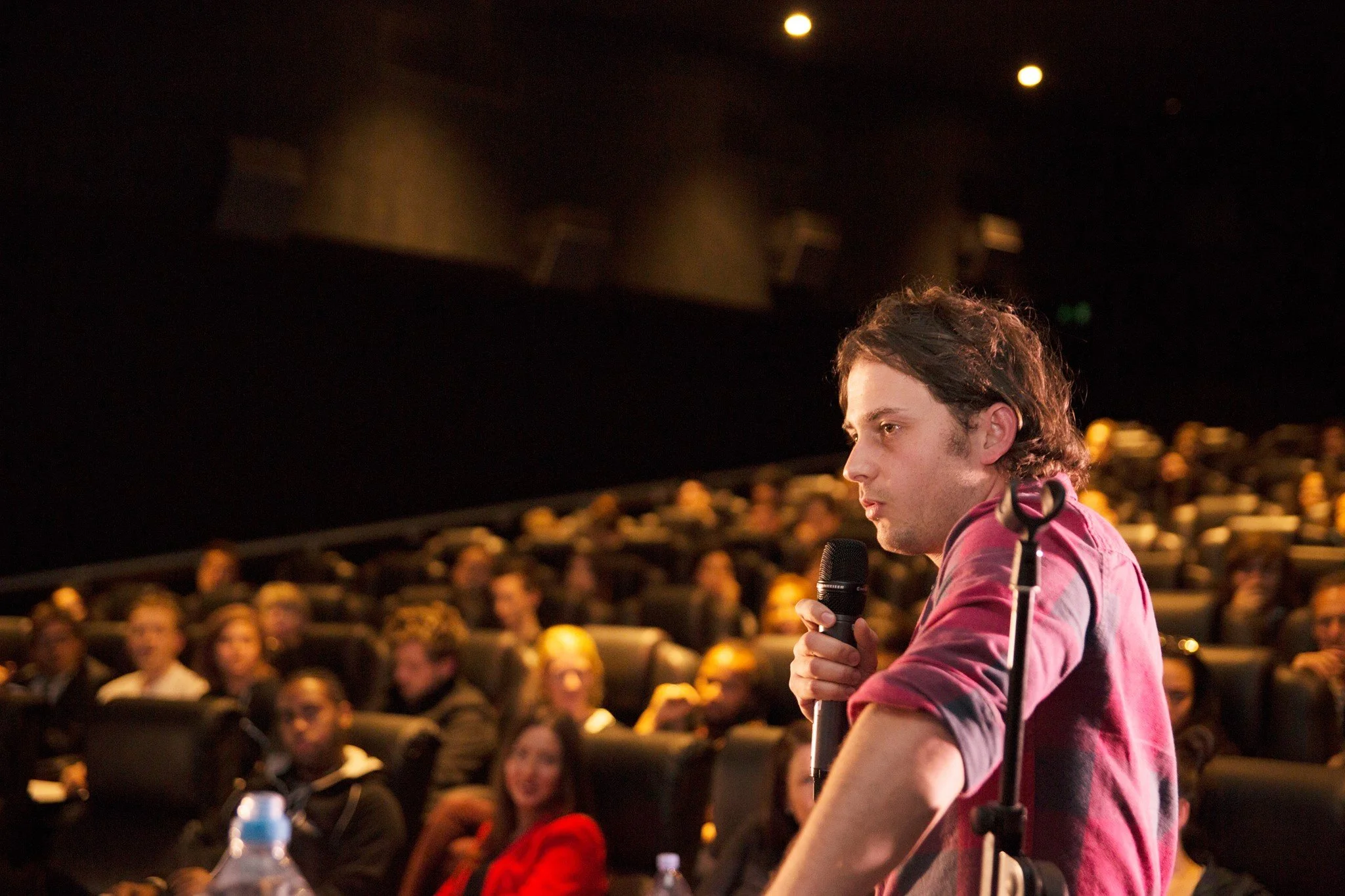 A young man in a red shirt speaking into a microphone at a conference or event, with an audience seated in the background in a dark auditorium.