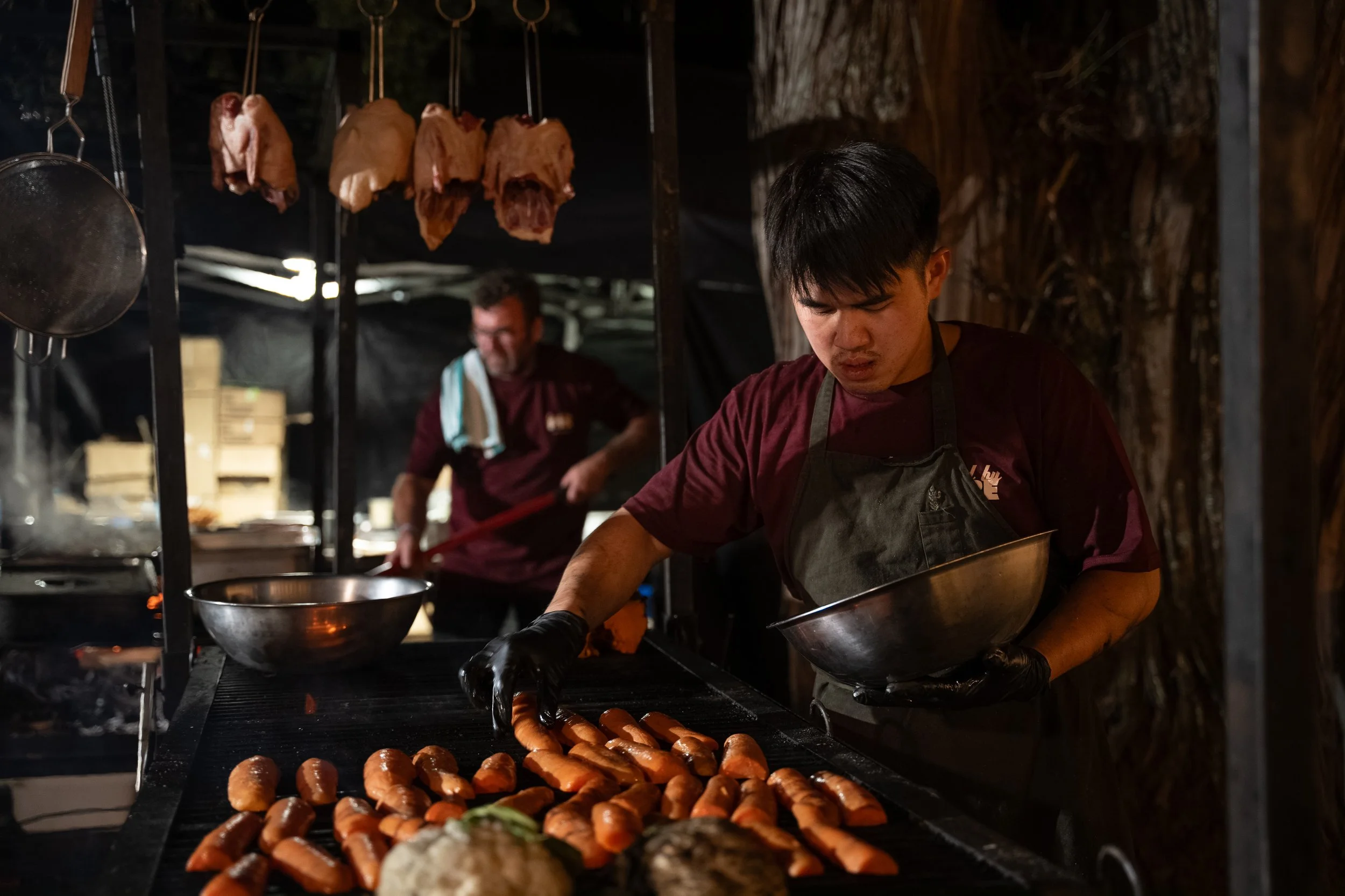 Two chefs preparing sausages on a grill at an outdoor food stand, with hanging meats in the background.