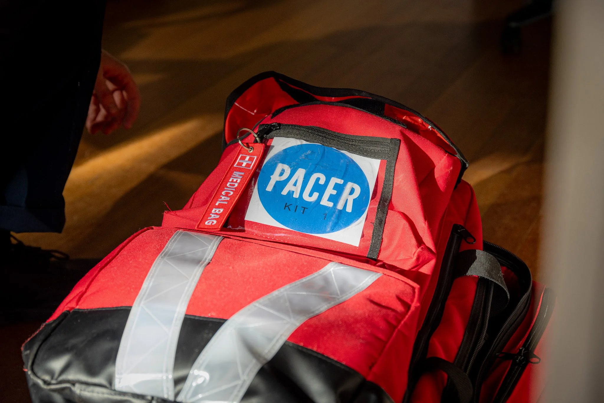 Red medical emergency kit bag with a blue sticker labeled 'PACER KIT 1' and a red tag that says 'MEDICAL BAG' resting on a wooden floor.
