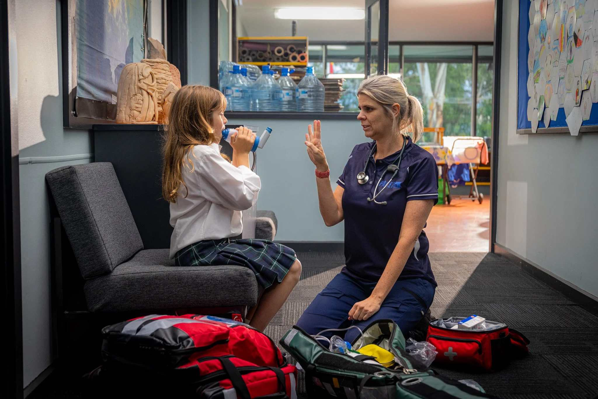 A female nurse wearing a dark blue uniform kneeling on the floor, speaking to a young girl in a school uniform who is sitting on a gray bench, using a medical device. The scene takes place in a brightly lit room with medical supplies and colorful dec