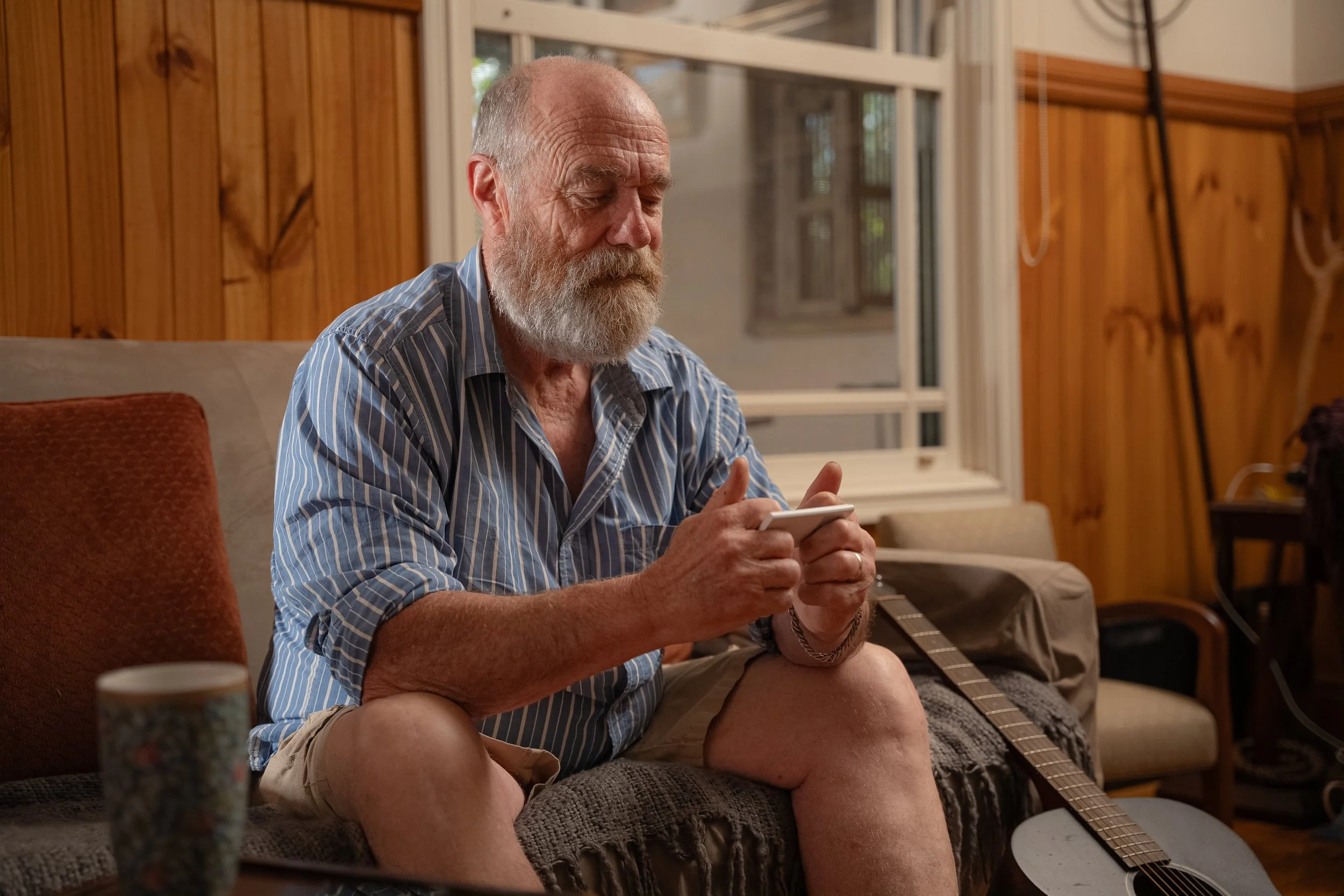 An older man with a gray beard and striped shirt sitting on a couch, looking at his phone, with a guitar resting on his lap in a cozy wooden-paneled room.