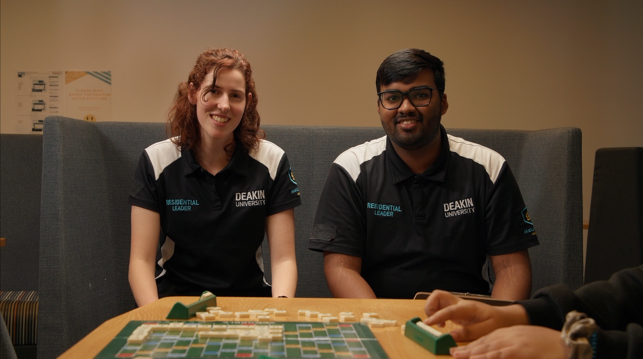 Two students sitting at a table playing a game of Mahjong, wearing black Deakin University shirts with a grey and beige background.