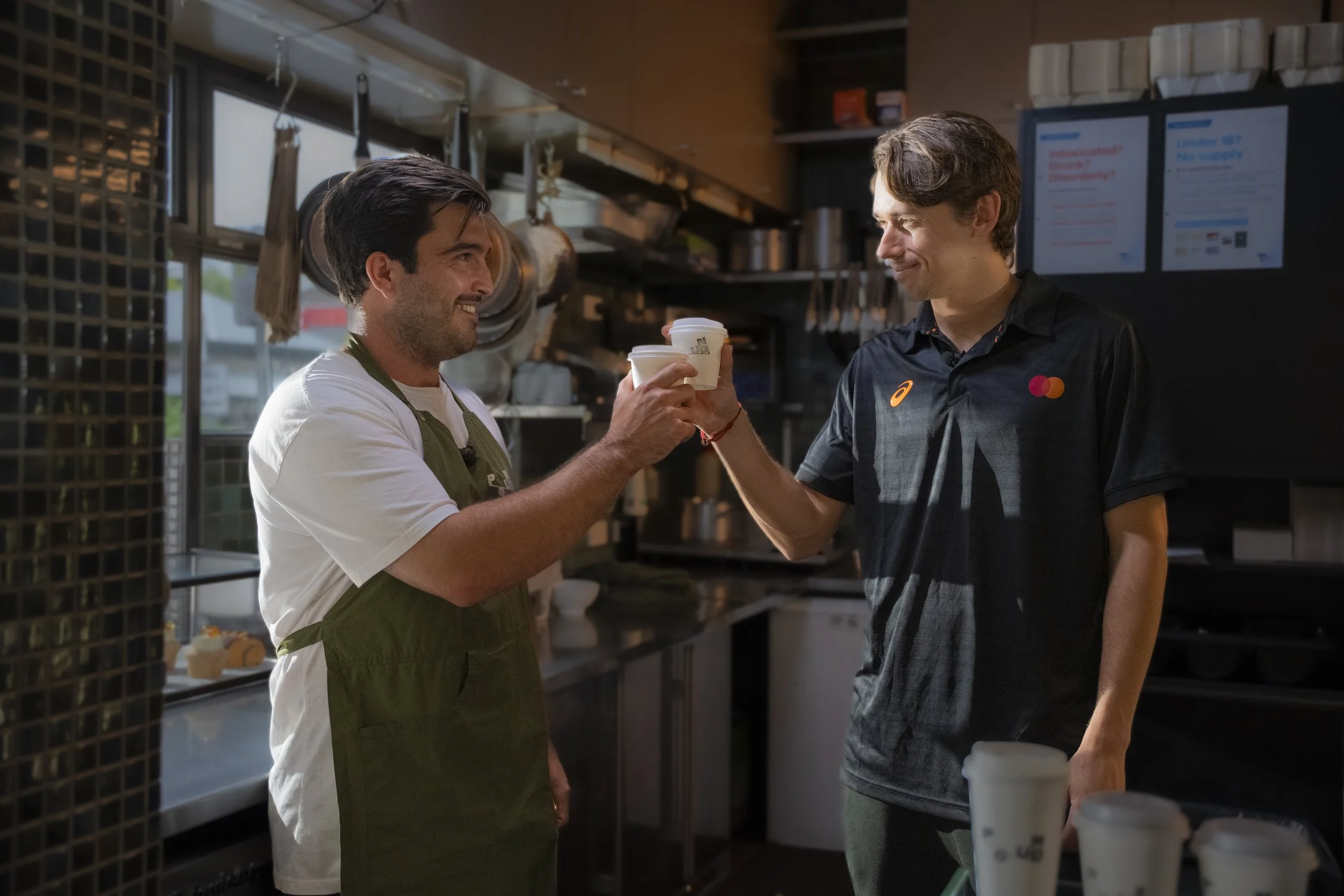 Two men in a kitchen clink paper cups, smiling at each other; one wears a white shirt with green apron, the other a black sports shirt with logos.