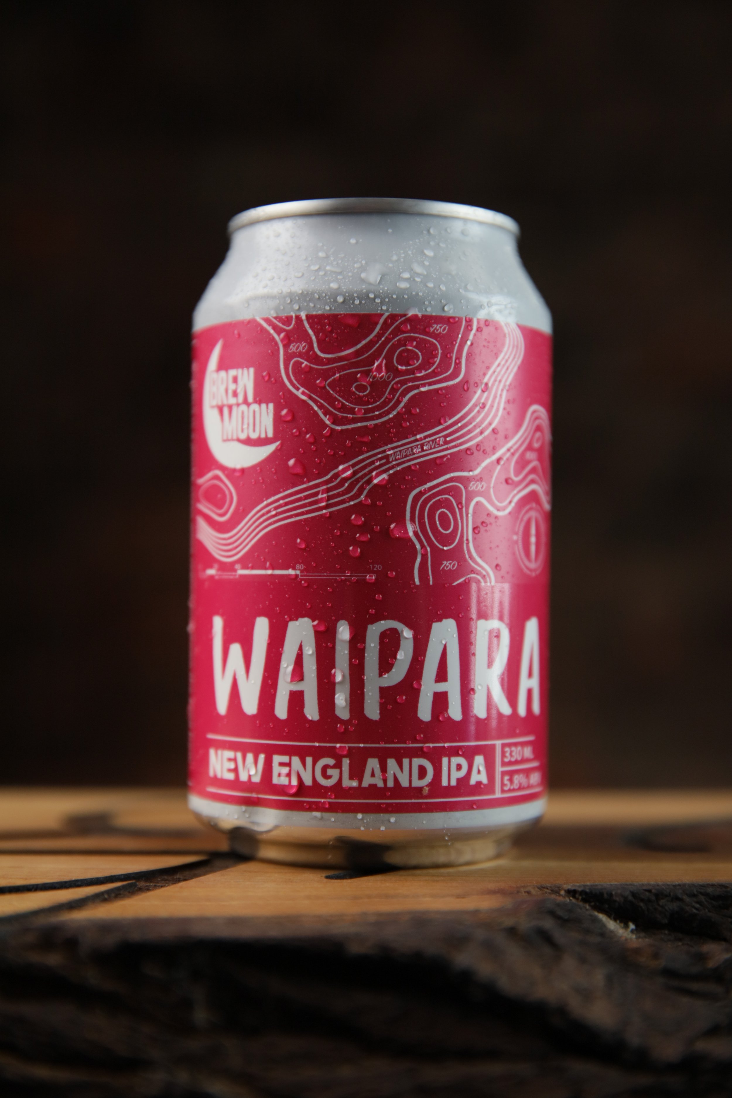 A pink can of Waipara New England IPA beer with water droplets on it, sitting on a wooden surface, with a dark, blurred background.