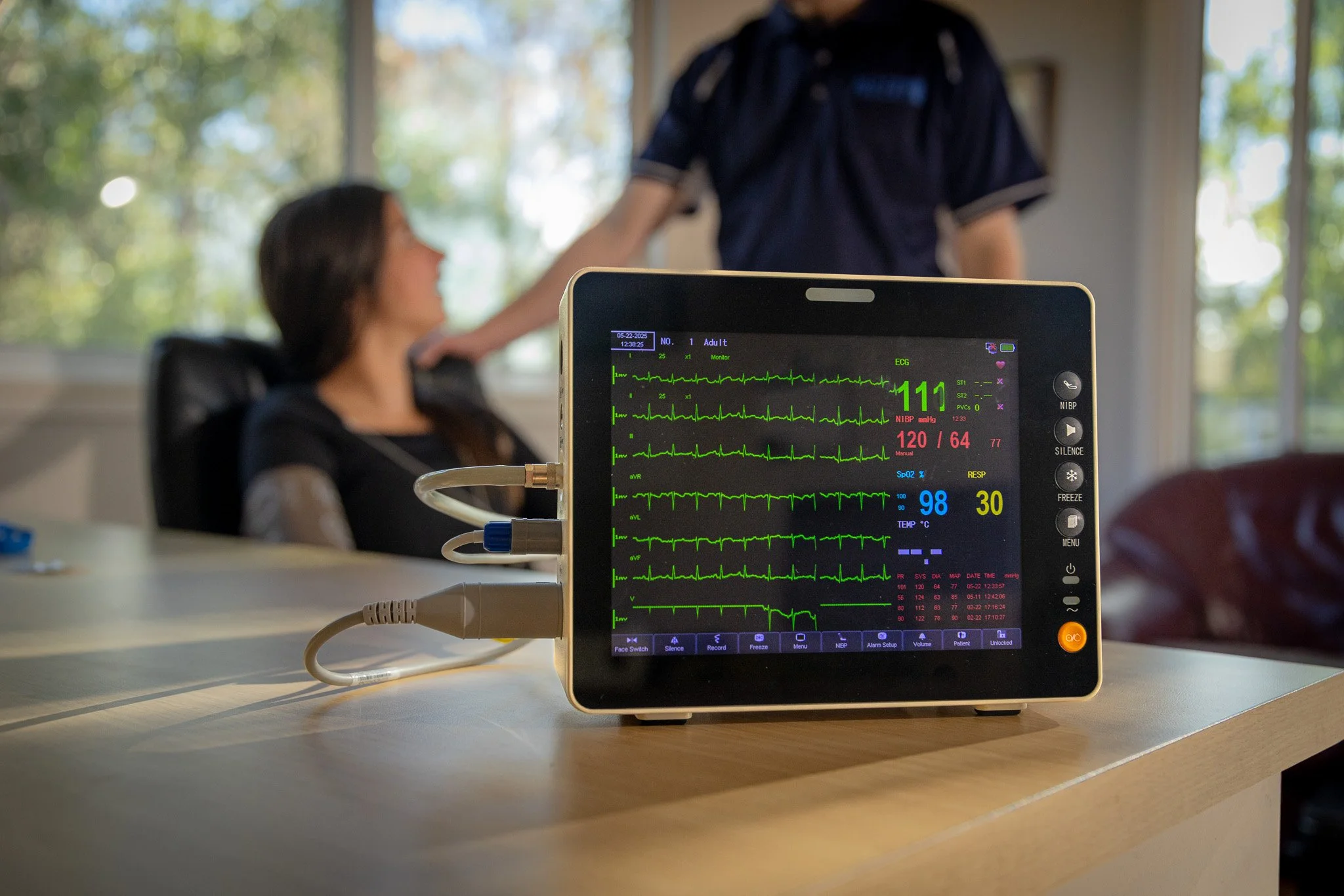 Medical monitor displaying patient vital signs such as heart rate, blood pressure, oxygen saturation, and temperature, with a healthcare provider and patient in the background.
