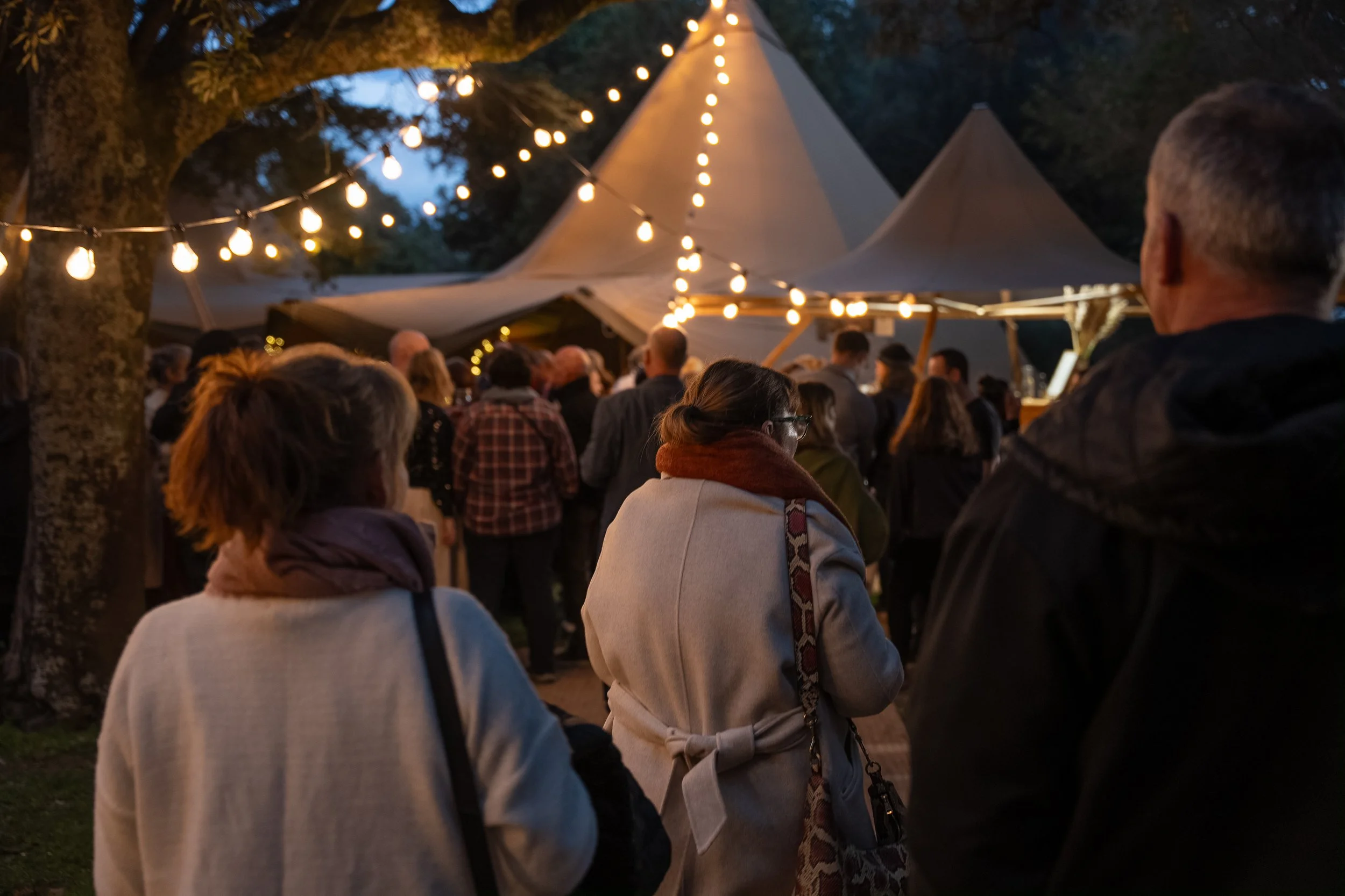 People gathered outdoors at night under string lights near tents or gazebos at an event or festival.