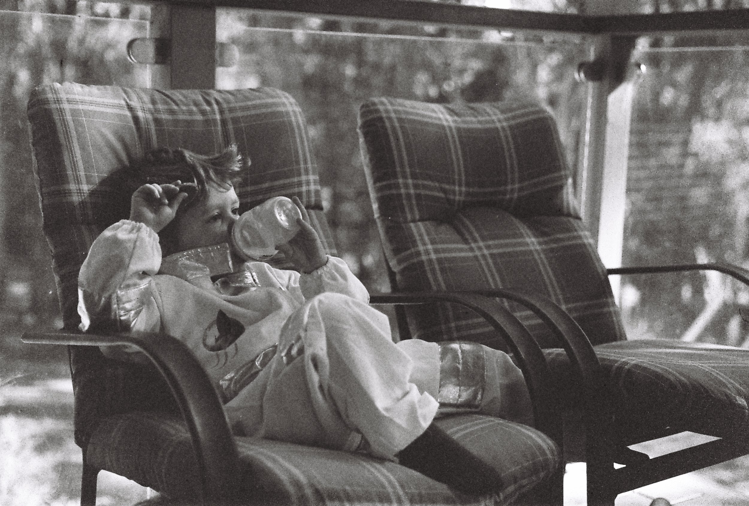A young girl with curly hair sipping from a bottle while relaxing on a plaid cushioned chair on a porch.