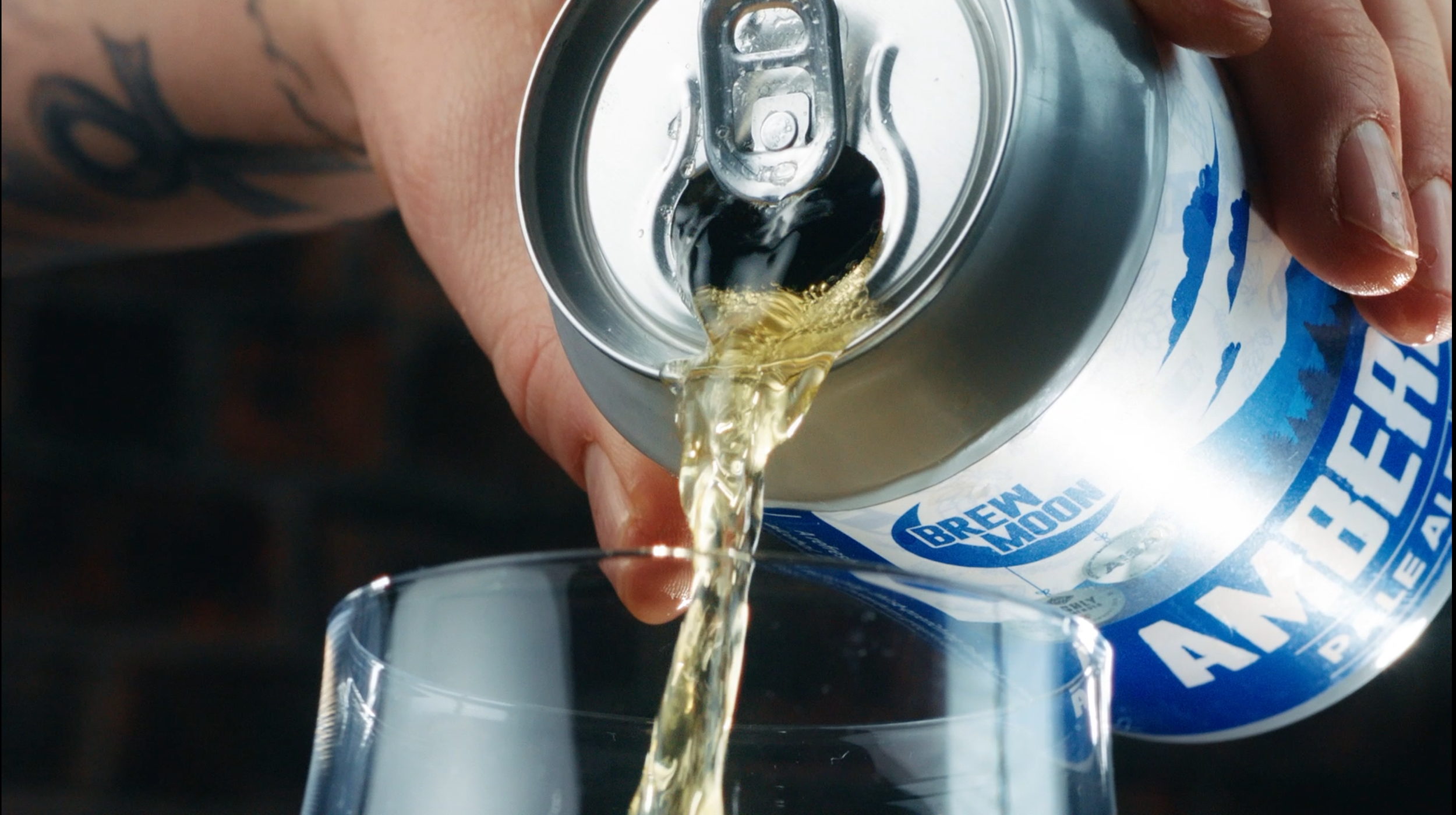 Person pouring light-colored craft beer from a can labeled 'Brew Moon' into a clear glass.