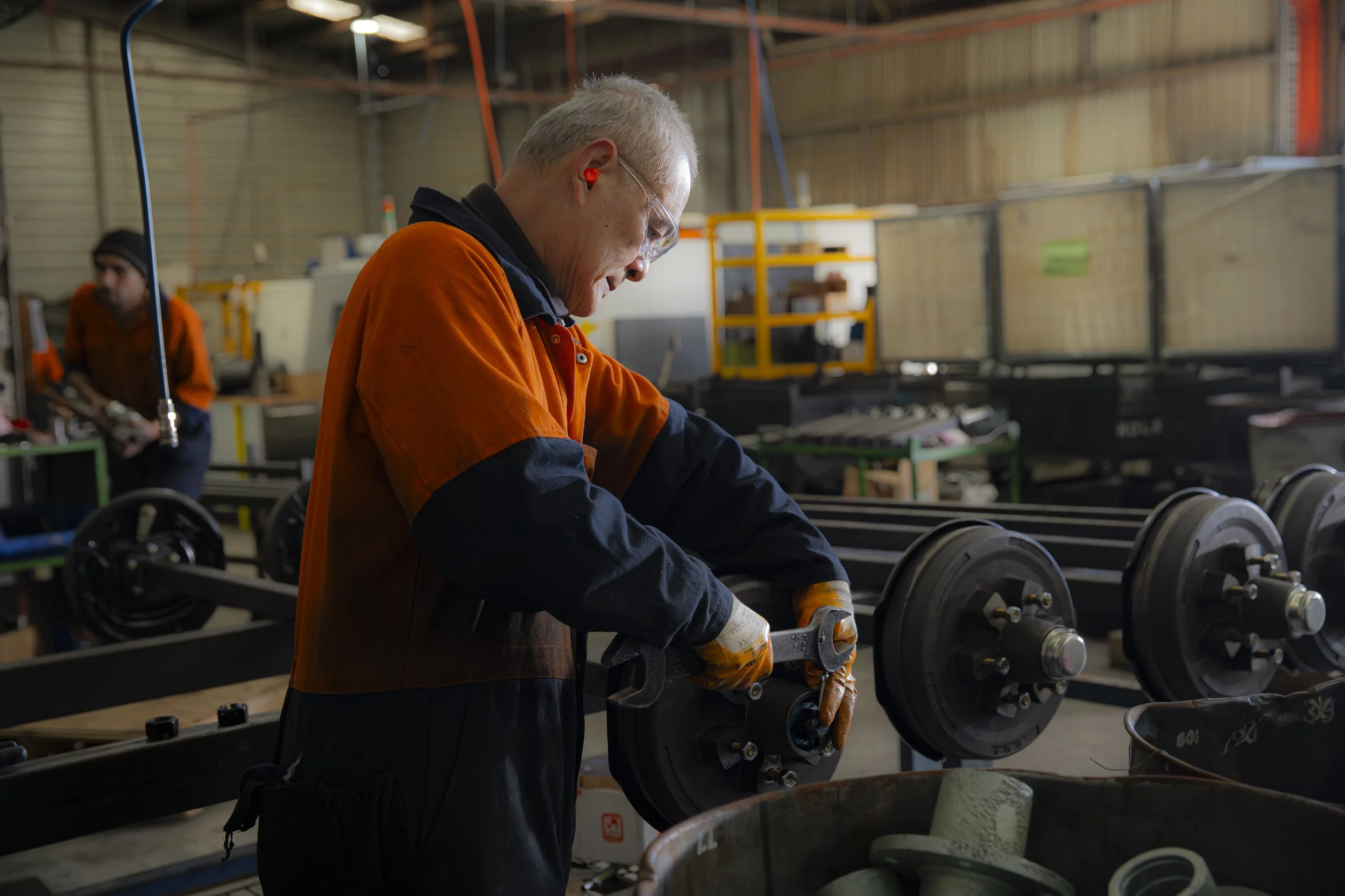 A worker in safety glasses and orange and black workwear inspects or repairs a wheel axle in an industrial workshop. Another worker is seen in the background, working on machinery.