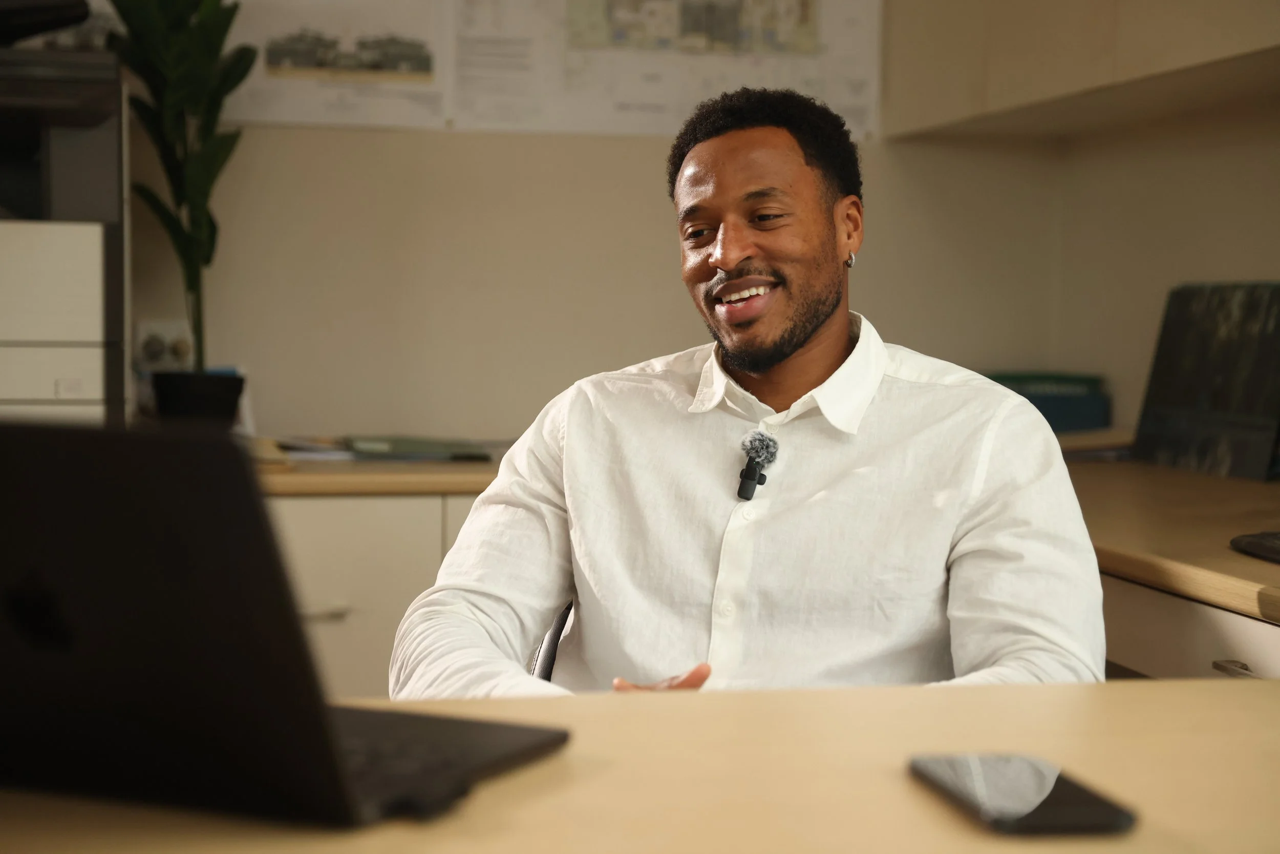 A man in a white shirt smiling while talking during a video call in an office setting.