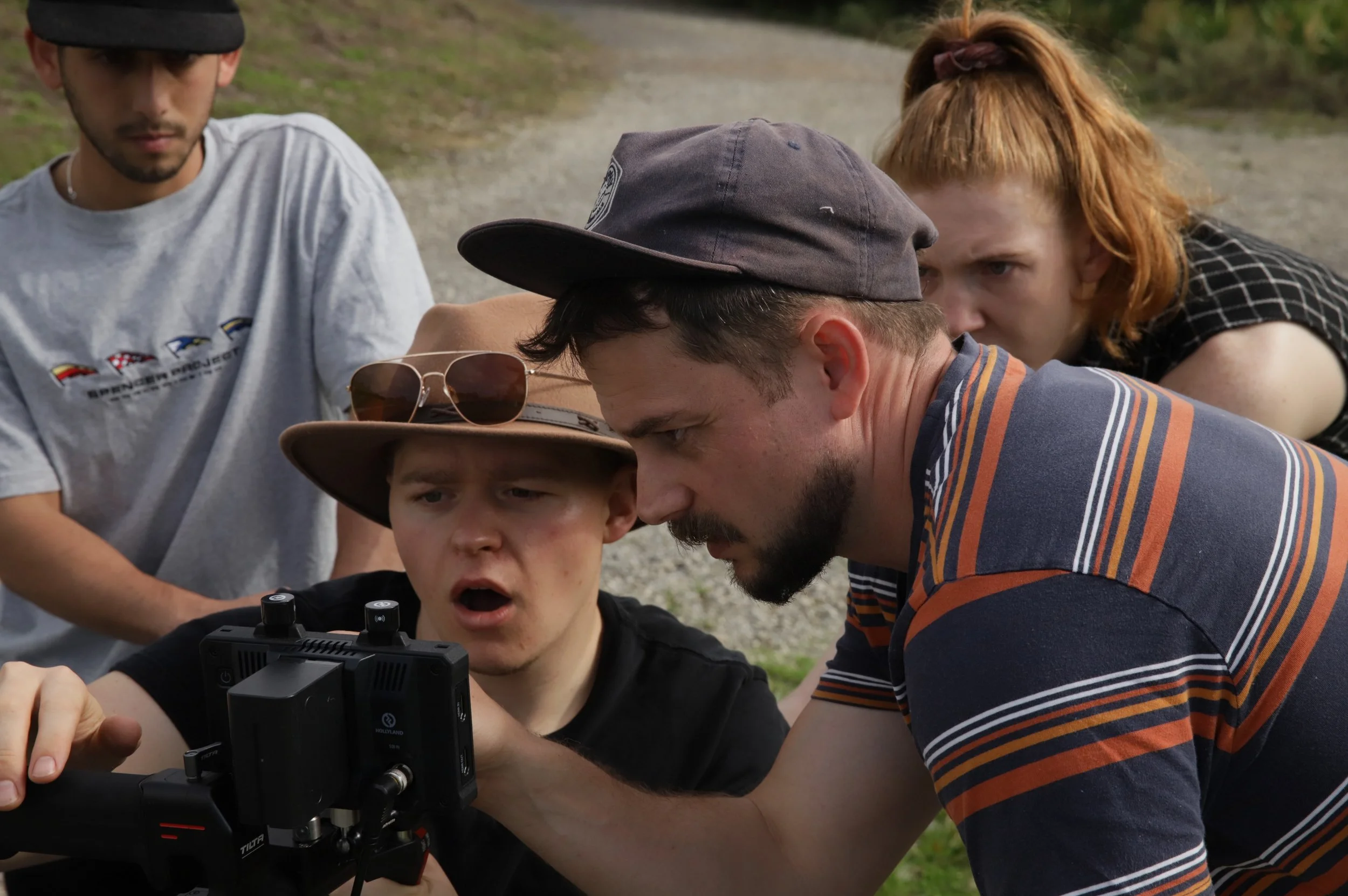A group of five people outdoors, gathered closely around a camera. One young man is operating the camera, with three others watching intently. The group appears to be working on a filming or photography project.