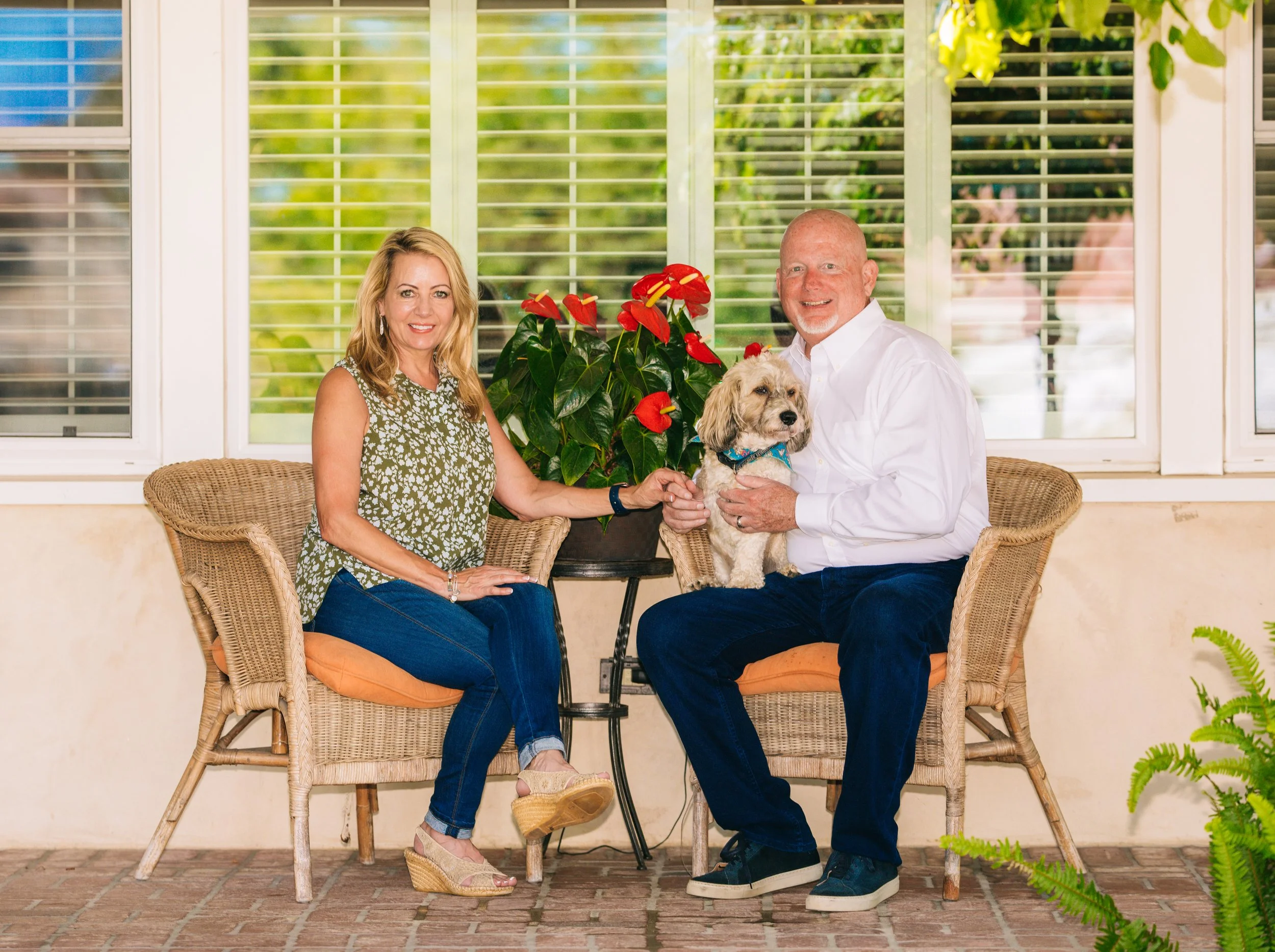 Mike Murchison, Candidate for Water Replenishment District, and his wife Tracy, outdoors on wicker chairs with a dog and red anthurium flowers, with window blinds and greenery in the background.