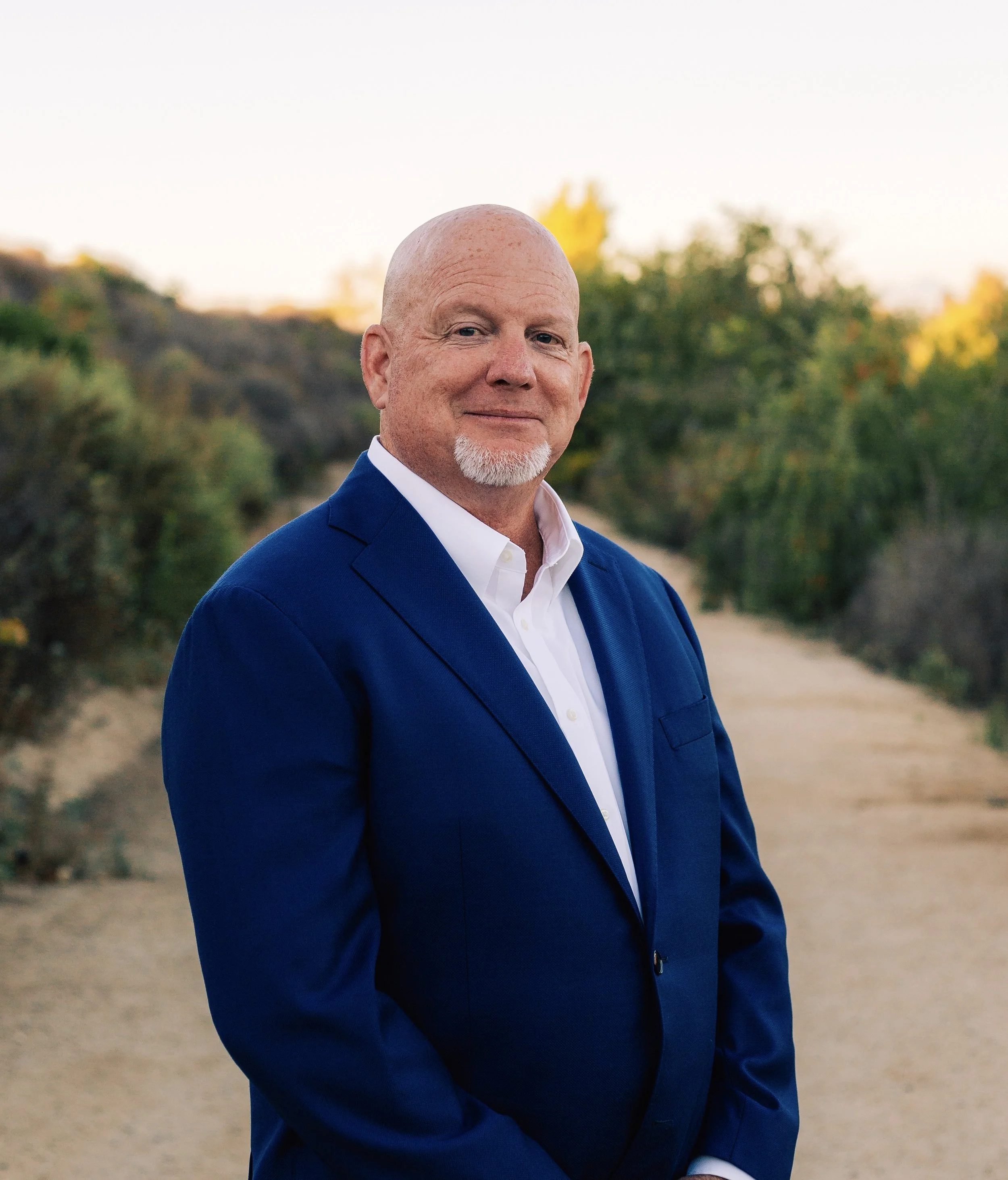 Mike Murchison, Candidate for Water Replenishment District, bald man with a goatee, wearing a blue suit and white shirt standing outdoors with trees and a trail in the background.