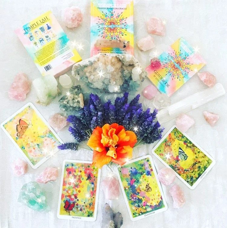 A colorful collection of tarot or oracle cards with butterflies and abstract art, surrounded by flowers, crystals, and stones on a light background.