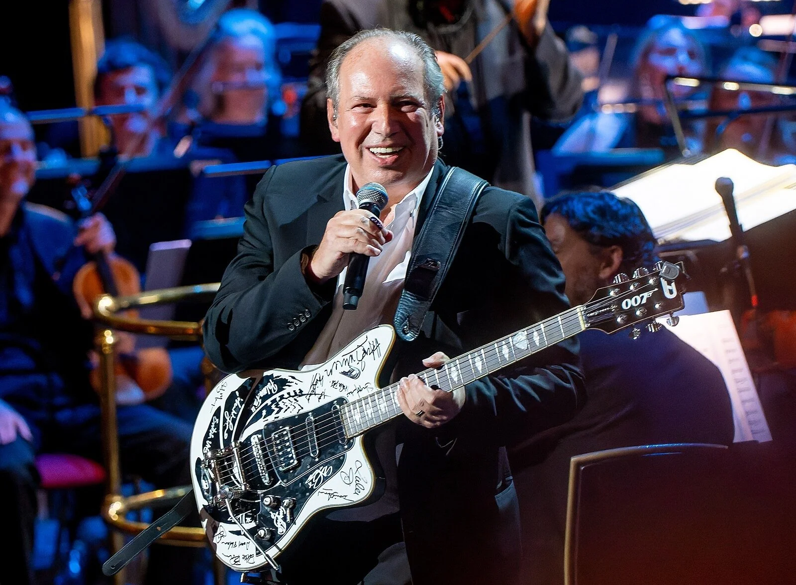 Hans Zimmer on stage with electric guitar and microphone. Orchestra is in the background.
