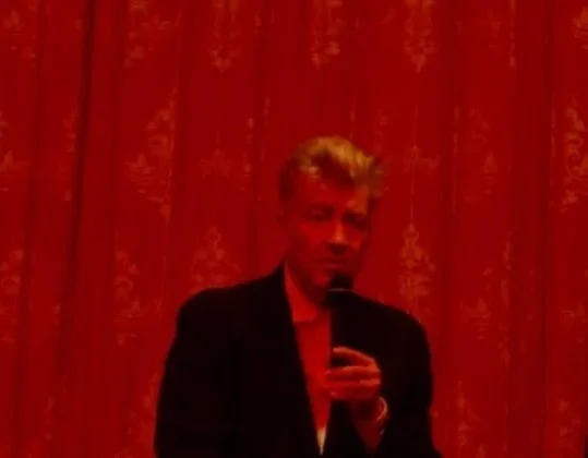 David Lynch with microphone in front of a velvet red curtain.