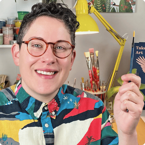 Steph (they/them) is wearing a colourful shirt, round glasses and holding a paintbrush in their hand. The background is their art studio.
