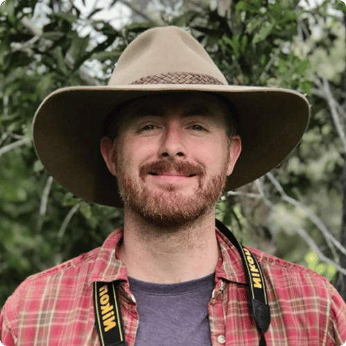 Ed is wearing a hat and flannel shirt. He has a camera around his neck.