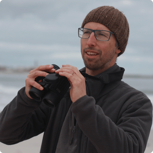 Golo wears long sleeves, a beanie and glasses and hold binoculars as he looks out into nature