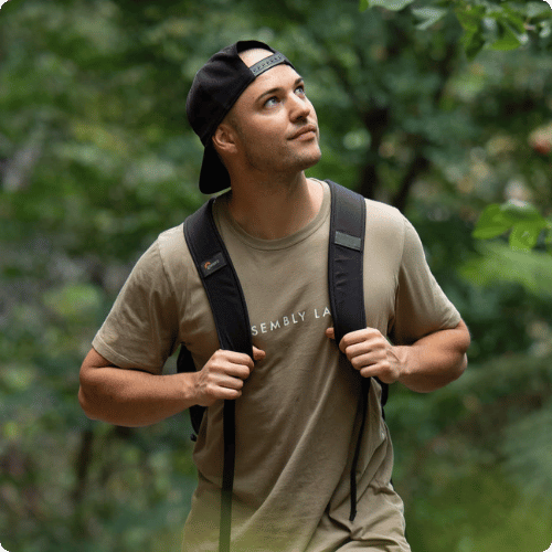 Jeremy (he/him) is wearing a backpack, has his cap on backwards and is staring up at the sky whilst walking in a forest