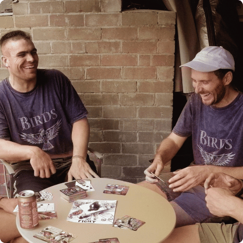 Chris and Corey (he/him) are sitting around a table with friends, playing their board game A Game Called Birds