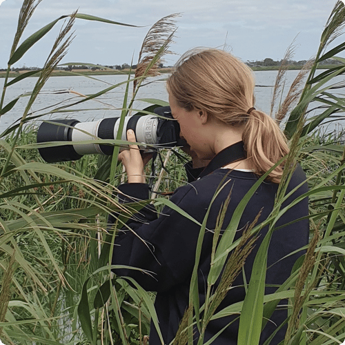 Georgia is standing amongst long grass looking into a zoom camera lens pointed towards a lake
