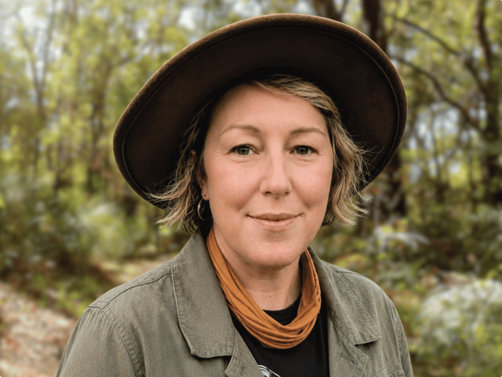 Kirsty is wearing a broad-brimmed hat, a mustard scarf and a green jacket. She is standing in the bush and smiling at the camera