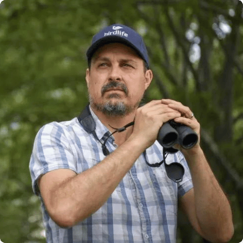 Sean is wearing a checkered shirt, birdlife australia hat and is holding binoculars
