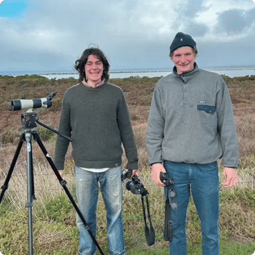 Cezary and Leo (he/him) are standing in a wetland. They have their camera on a tripod and are both holding binoculars.