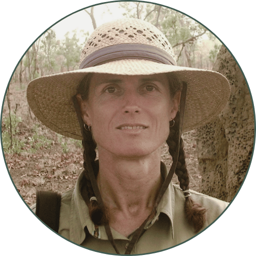 Ricki (she/her) is wearing a straw hat and green shirt, standing in front of a large termite mound in the bush