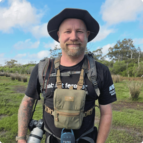 Ben (he/him) is smiling at the camera and wearing a Trust for Nature t-shirt. Around his body is a camera, pair of binoculars and a small bag strapped to his chest.