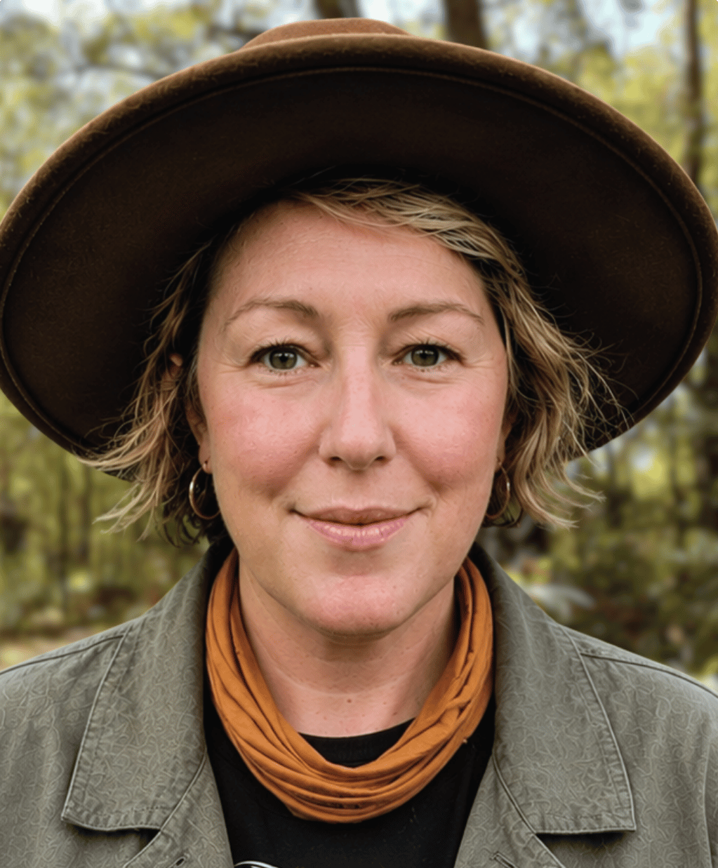 Kirsty is wearing a broad-rimmed hat, a mustard scarf and green jacket. She is smiling at the camera and standing in a forest