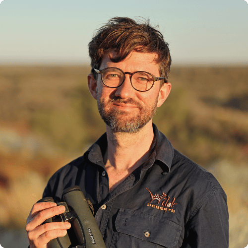 Tom has short brown hair and a beard, he stands looking at the camera with a pair of binoculars in his hands