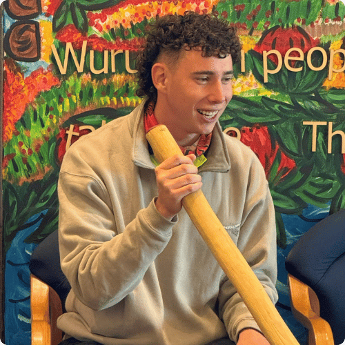 Kai (he/him) is sitting on a chair and smiling at the audience
