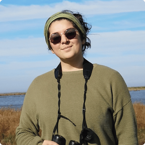 Kristal stands in front of a lake in a green jumper and matching headband with binoculars around her neck and sunglasses on her face