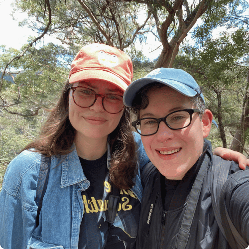 Stephanie and Amy stand together surrounded by nature, both wearing caps and glasses