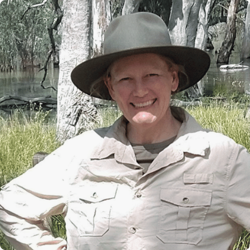 Lareen smiles at the camera wearing a wide brim hat and a long sleeve shirt, she stands in front of a wetland area