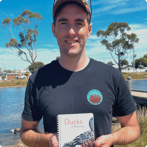 Jason is standing in front of a pond and holding his book Ducks of Tasmania
