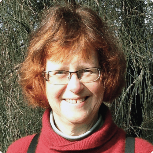 Ann has short hair with a fringe and wears glasses, she is smiling at the camera