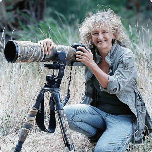Kim kneels on the ground next to a camera on a tripod with a long lens, she has curly blonde hair and wears casual clothes