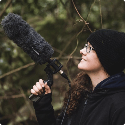 Vicky is wearing a black jumper and beanie and holds a large microphone and looks up into the trees