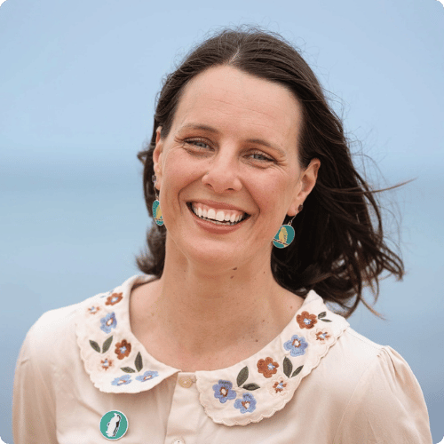 Paula (she/her) is smiling at the camera. She is wearing a blouse with a flower printed collar and bird earrings