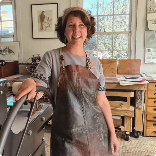 Bridget stands with a grey shift and apron over the top in a painting studio, she ha short dark hair and wears a headband