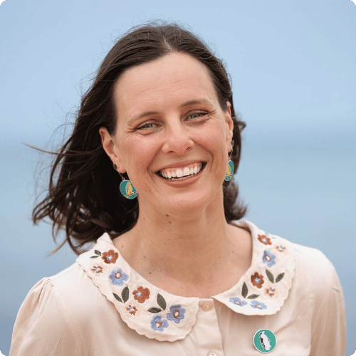 Paula (she/her) is wearing a floral blouse and budgie earrings